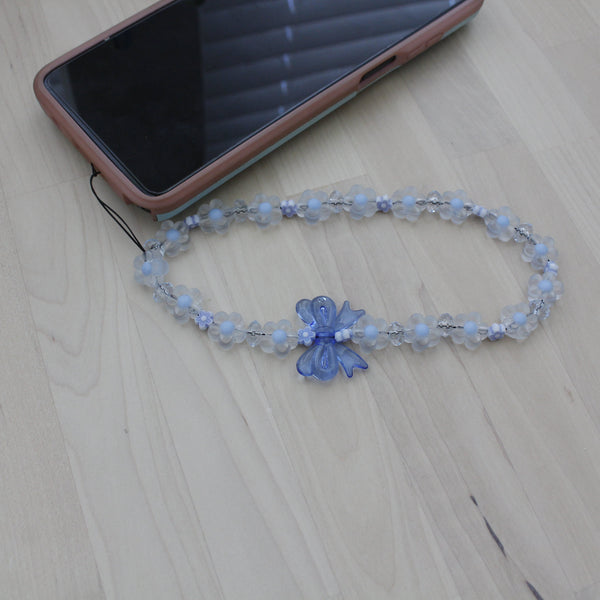 Pastel blue flower phone strap with bow displayed beside a smartphone on a wooden surface.