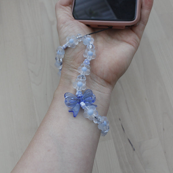 Pastel blue flower phone strap with bow worn on wrist, holding a smartphone, ideal for women and girls.