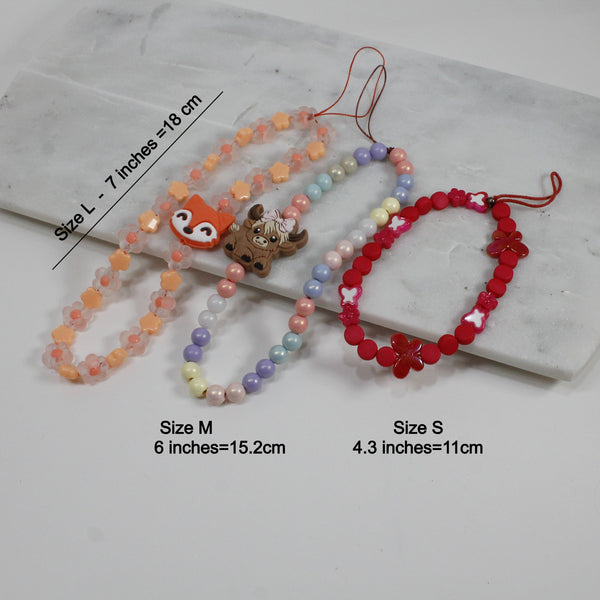 Three colorful phone straps laid out on marble: size S (11cm), M (15.2cm), and L (18cm) with fun designs and beads.