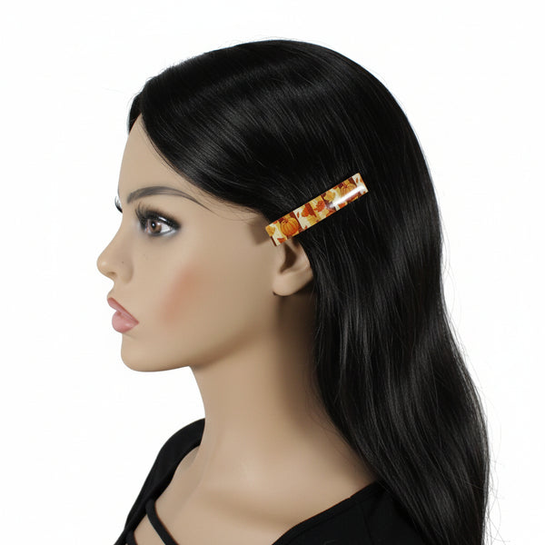 A side-profile view of a mannequin wearing a long black wig with a rectangular barrette hair clip featuring a warm-toned autumn leaf pattern in orange, yellow, and brown on a white background.