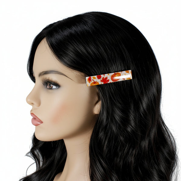A side-profile view of a mannequin head with long black hair, wearing a slim rectangular hair clip decorated with an autumn pattern of orange pumpkins and red leaves on a white background.