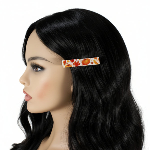 A close-up side view of a mannequin head with long black hair, wearing a slim rectangular hair clip decorated with an autumn leaf and pumpkin pattern on a white background.