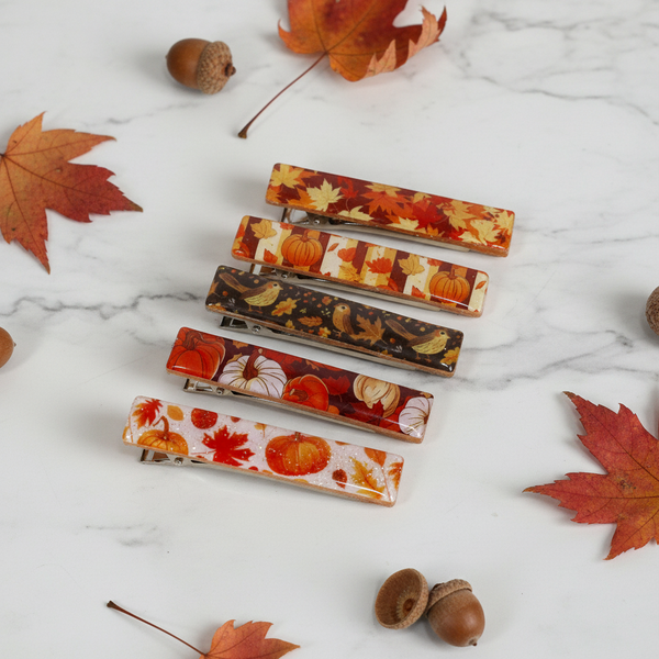 Five hair claw clips arranged diagonally on a white background, featuring various autumn-themed patterns including pumpkins, maple leaves, and fall foliage.