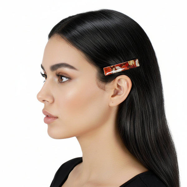 A side-profile view of a mannequin head with long black hair and a small rectangular hair clip holding a section of hair. The clip has a red-orange background with a floral pattern in white, yellow, and green.