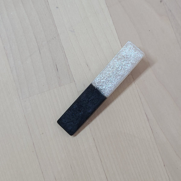 A rectangular makeup sponge with a dual-tone finish, featuring a textured black bottom half and a glittery silver top half, resting on a light wood grain surface.