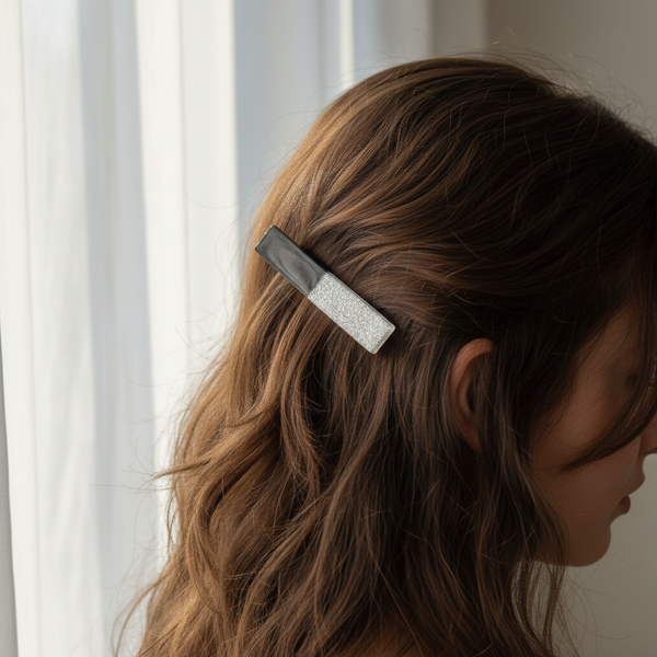 A woman with wavy brown hair wearing a rectangular hair clip with a black and white glitter design, shown in profile against a soft window background.