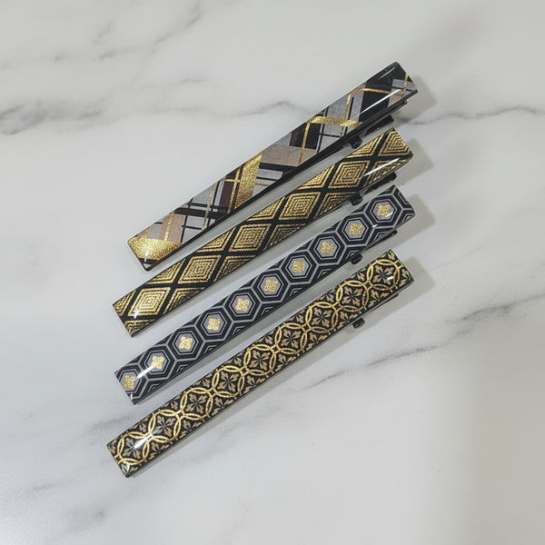 Four slim rectangular accessories with decorative geometric patterns in black, gold, and beige, arranged diagonally on a plain light background.