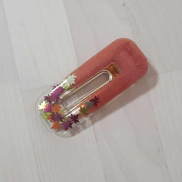 A clear plastic hair clip filled with shimmering peach-pink glitter and decorated with colorful metallic maple leaf confetti, resting on a white wooden surface.