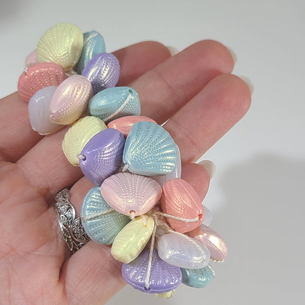 A close-up of a hand holding a scrunchie adorned with pastel seashell-shaped beads in soft pink, lavender, light blue, and pale yellow.
