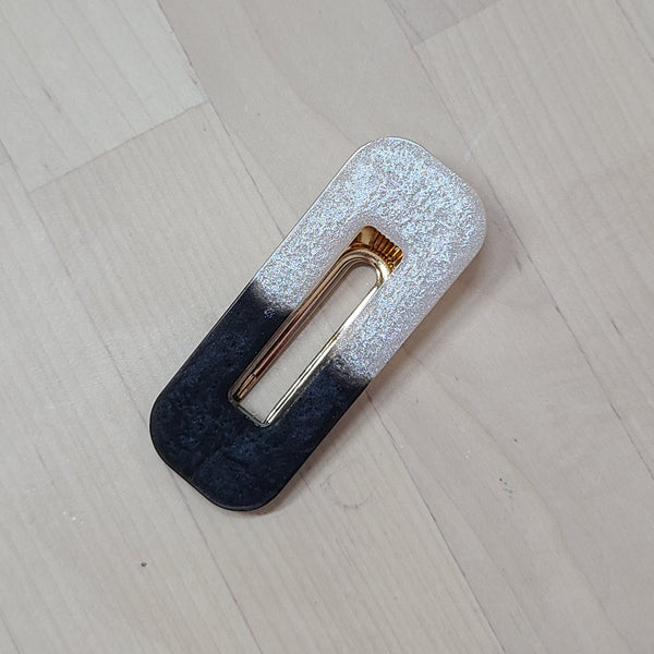 A rectangular hair claw clip with a two-tone design, featuring a glittery silver top half and a dark navy blue bottom half, with a gold metal spring and clip mechanism in the center, resting on a light wood floor.