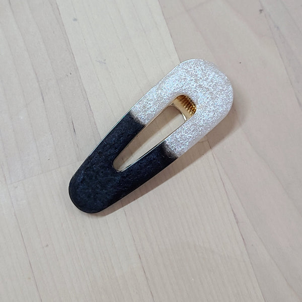 A split-design hair clip with a textured black and glittery silver finish, resting on a light wood grain surface.