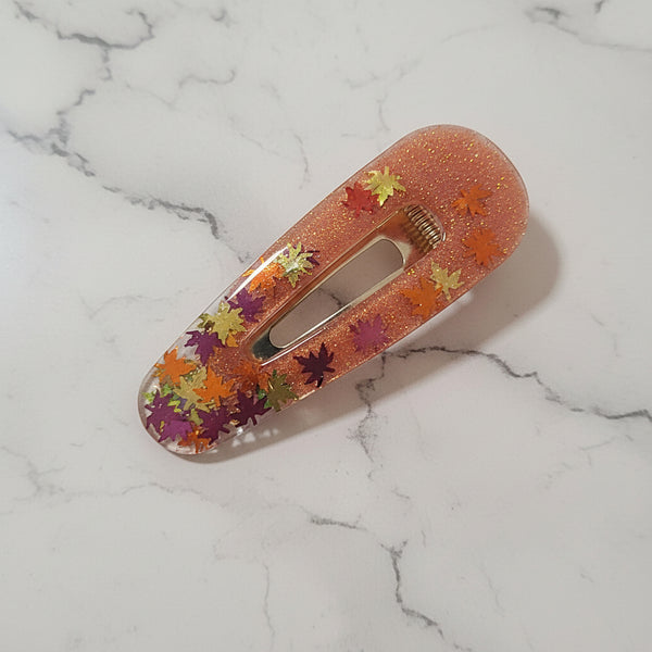 A clear acrylic hair clip filled with shimmering peach-pink glitter and decorated with colorful dried autumn leaves, shown on a light wood background.
