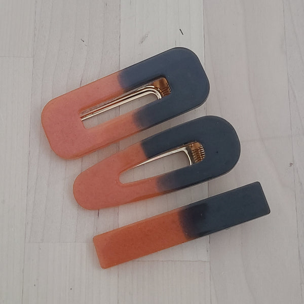 Three hair claw clips with a dual-tone gradient design transitioning from terracotta orange to dark navy blue, arranged diagonally on a light-colored wooden surface.